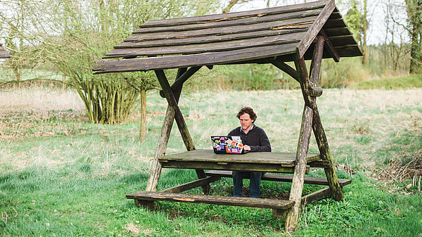 Foto: Eine Person sitzt mit einem Laptop an einem überdachten Picknicktisch.