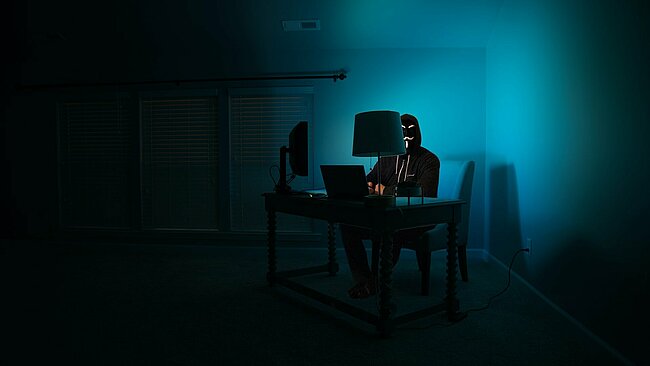 Ein Mann an einem Computer, der als anonymer Hacker verkleidet ist und eine Guy-Fawkes-Maske trägt.